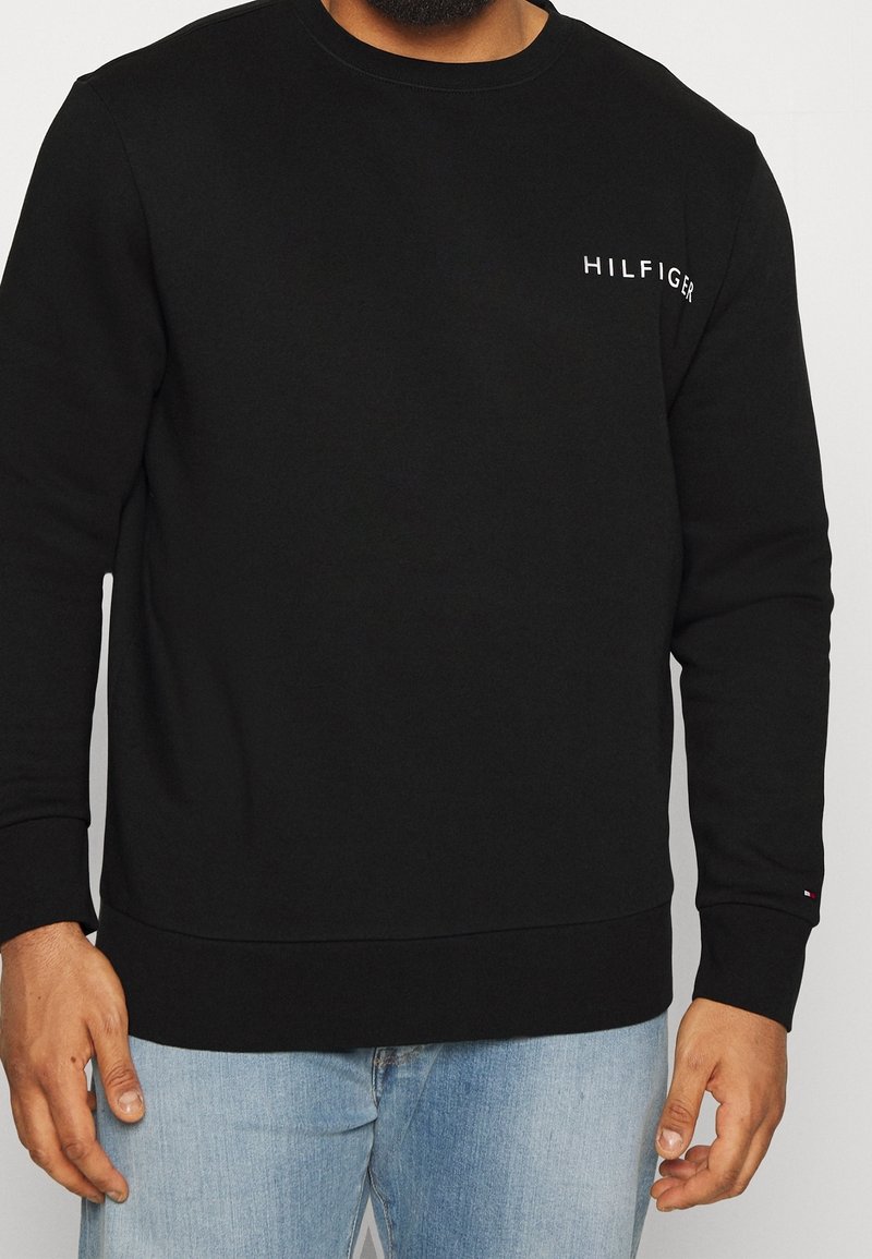 Homme portant un sweat-shirt noir à manches longues avec le texte « HILFIGER » sur la poitrine et un petit logo sur le poignet de la manche gauche, associé à un jean bleu clair.