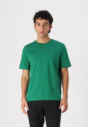 Tânăr cu păr scurt și creț purtând un tricou verde cu mâneci scurte și pantaloni negri, stând în fața unui fundal alb simplu.