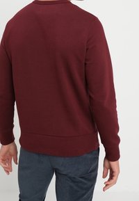 Felpa bordeaux con maniche lunghe, coltondo, polsini e orlo a costine, abbinata a pantaloni scuri strutturati, vista da dietro.