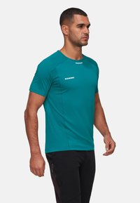 Man som bär en teal Mammut t-shirt med kort ärm och svarta byxor, stående på sidan mot en enkel ljus bakgrund.