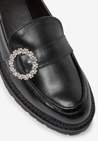 Schwarze Lederloafer mit horizontalen Bändern und einem runden Strassakzent. Glatte Textur, abgerundete Zehen und Gummisohle mit Nähten.