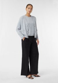 Hellgrauer Cardigan mit Knöpfen, getragen über einem passenden Pullover. Schwarze weit geschnittene Hose und schwarze Riemchen-Sandalen runden das Outfit ab.