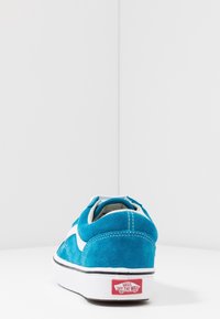 Modrý semišový střevíc s bílými akcenty, šněrováním, plochou podrážkou a červeným logem na patě. Texturovaný materiál a zakřivený tvar.