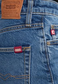 Jeans iz žameta v srednji modri barvi z kontrastnim rumenim šivom, z rdečimi tkani oznakami na žepih in rjavo usnjeno oznako na pasu.