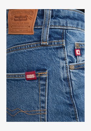 Keskmise sinise denimpüksid, millel on kontrastsete kollaste õmblustega, taskutes punased kudumislabelid ja seljal pruun nahast logo tempel.