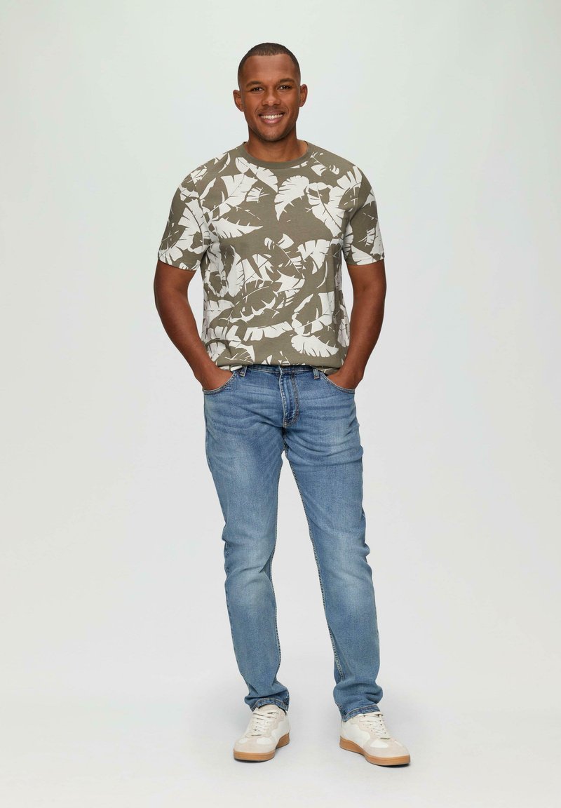 Camiseta verde oliva de manga corta con patrón de hojas blancas, combinada con jeans azul claro y zapatillas blancas. El modelo está de pie con las manos en las caderas.