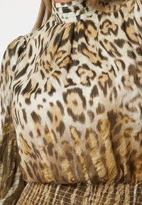 Blouse à imprimé léopard avec encolure froncée, manches longues bouffantes et tissu texturé aux nuances de beige, marron et accents dorés.