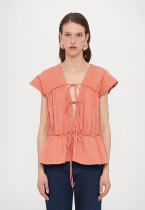 Peach-colored top with cap sleeves, gathered texture, deep V-neck, and adjustable drawstring at the waist. Paired with blue jeans.