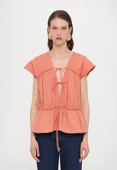 Top di colore pesca con maniche a campana, texture arricciata, scollatura a V profonda e coulisse regolabile in vita. Abbinato a jeans blu.