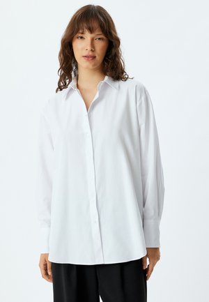 Camicia bianca con maniche lunghe, colletto e taglio dritto. Tessuto liscio, con una leggera lucentezza e chiusura frontale a bottoni.
