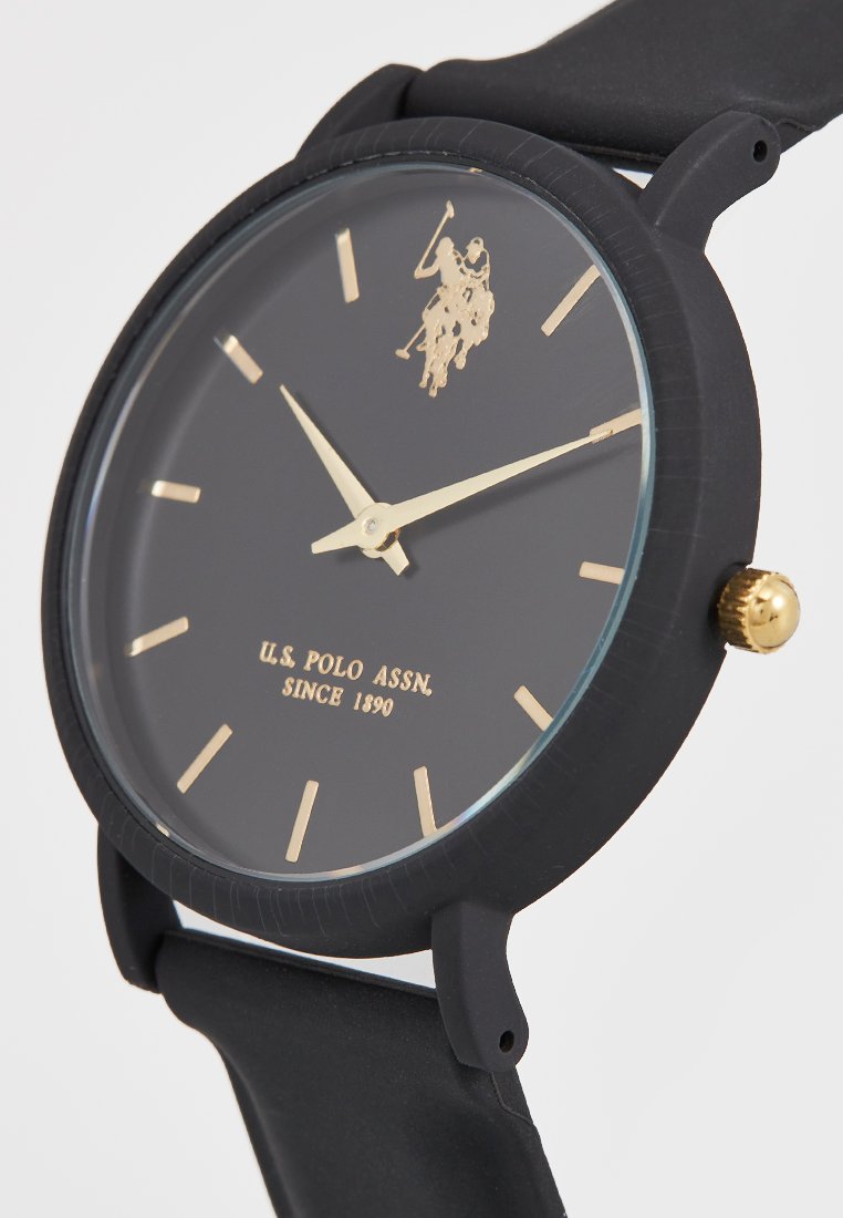 Reloj de silicona negra con una esfera monocromática y acentos dorados, un logo dorado y marcadores de hora minimalistas. Diseño de caja redondeada.
