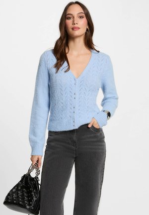 Cárdigan de punto azul claro con cuello en V, patrón de encaje intrincado y botones plateados. Combinado con jeans negros y un bolso negro.