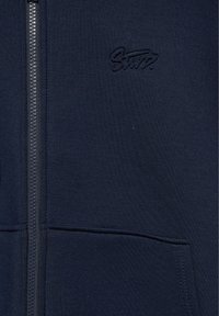 Sweat à capuche zippé bleu foncé avec une poche avant et un petit logo brodé sur la poitrine.
