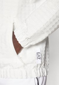 Biela textúrovaná bunda s vreckom na zips, elastickými manžetami a potlačenou logom. Materiál má vystúpený vzor.