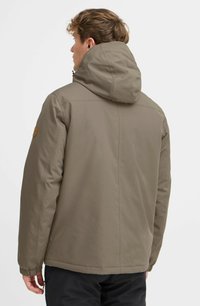 Chaqueta marrón con capucha, de textura suave y puños elásticos. Presenta una capucha redondeada y un parche con el logo en el hombro izquierdo.