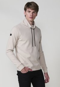 Giovane uomo che indossa un pullover beige a trama con lacci neri e pantaloni neri, in piedi con una mano in tasca su uno sfondo neutro.