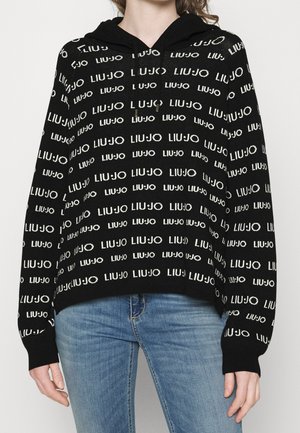 Osoba v černé mikině s kapucí s bílým vzorem textu „LIU JO“ a modrými džínami, ruce volně podél těla.