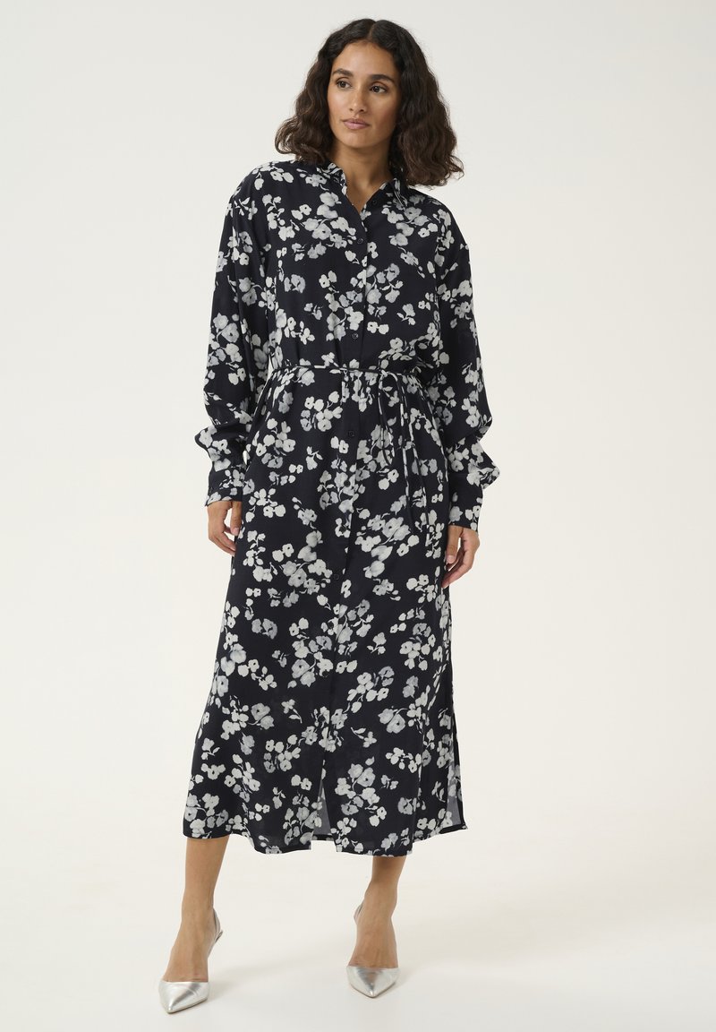 Robe midi florale avec un fond bleu marine foncé, dotée de motifs floraux blancs, de manches longues, d'une fermeture à boutons et d'une taille nouée. Texture de tissu lisse.