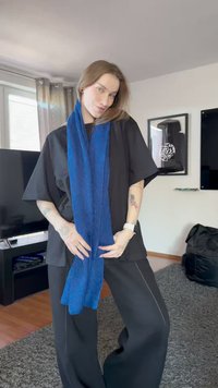 Jaunas žmogus su tatuiruotėmis, apsirengęs juodu kostiumu ir mėlynu šaliku, stovi svetainėje su televizorium ir rėminiais meno kūriniais.