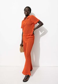 Ensemble en deux pièces tricoté orange avec un ourlet festonné, comprenant un haut polo à manches courtes et un pantalon évasé. Accents de sac en osier.
