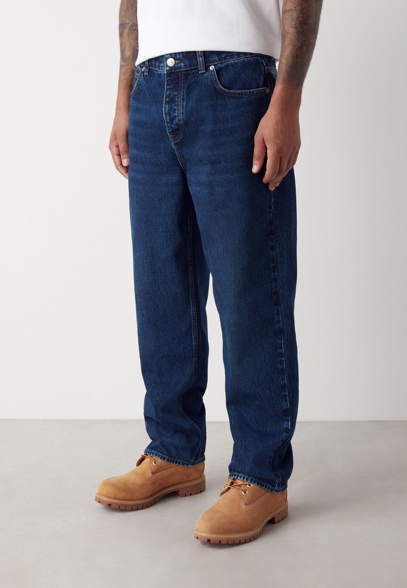 Muž v tmavomodrých rovno strihaných džínsoch, bielej košeli, hnedej pracovnej obuvi na zaväzovanie a s viditeľnými tetovaniami na predlaktiach.