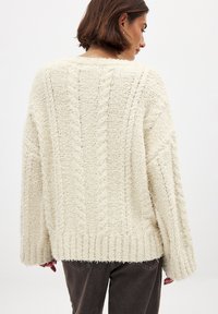 White, textured knit sweater with a loose fit, featuring a ribbed hem and cable-knit pattern; worn over dark trousers.