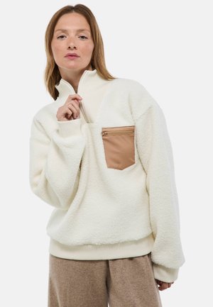 Pull en polaire blanc avec un col montant, présentant une poche zippée beige sur la poitrine, des poignets côtelés et un ourlet pour un ajustement confortable.