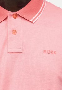 Κοντινό πλάνο ενός ροζ πόλο με δύο κουμπιά, λευκές διπλές ρίγες στο γιακά και το λογότυπο "BOSS" στο στήθος.