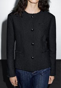 Žena v černém strukturovaném sako na knoflíky s dlouhými rukávy a tmavě modrých džínách, stojící na jednobarevném pozadí.