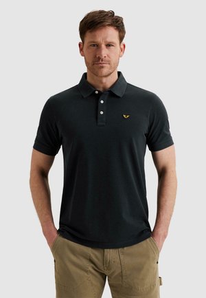 Hombre con cabello corto castaño, vistiendo una camiseta polo negra con tres botones blancos y pantalones beige, con las manos en los bolsillos.