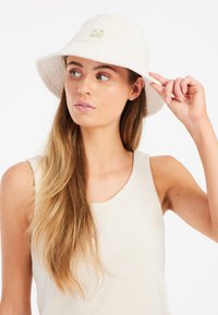 Witte badstof bucket hat met een brede rand en subtiele vouwdetails, voorzien van een klein gouden logo op de voorkant. Zachte textuur.