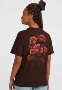 T-shirt en coton marron avec un grand motif floral en rose et orange au dos, agrémenté d'accents de feuillage et d'éléments graphiques de texte.