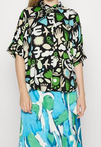 Blusa a maniche corte realizzata in tessuto leggero, con uno sfondo nero e audaci forme astratte nei colori verde, blu e crema.