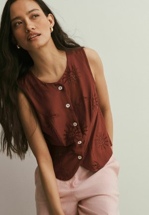 Frau trägt ein ärmelloses braunes Hemd mit Knöpfen und Sonnenstickerei sowie hellrosa Hose, schaut leicht nach oben und zur Seite.