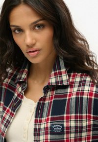 Camicia di flanella a quadri rossi, blu navy e bianchi con colletto, abbinata a una camicia color crema con bottoni. Presenta un’etichetta del marchio sulla camicia.