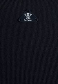 Černá pletená látka s ribbovanou texturou, na které se nachází trojúhelníková reliéfní nášivka s nápisem "Barbour" a rokem "1894" v bílé barvě.