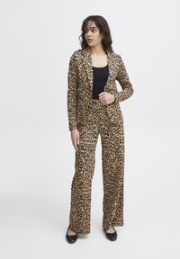 Completo giacca e pantaloni con stampa leopardata in beige e nero; design strutturato con maniche lunghe, abbinato a una maglietta nera e scarpe basse.