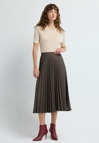 Atasan lengan pendek berwarna krem dengan kerah yang dihias dipadukan dengan rok panjang berlipat berwarna coklat tua dan sepatu bot pergelangan kaki hak tinggi berwarna burgundy.