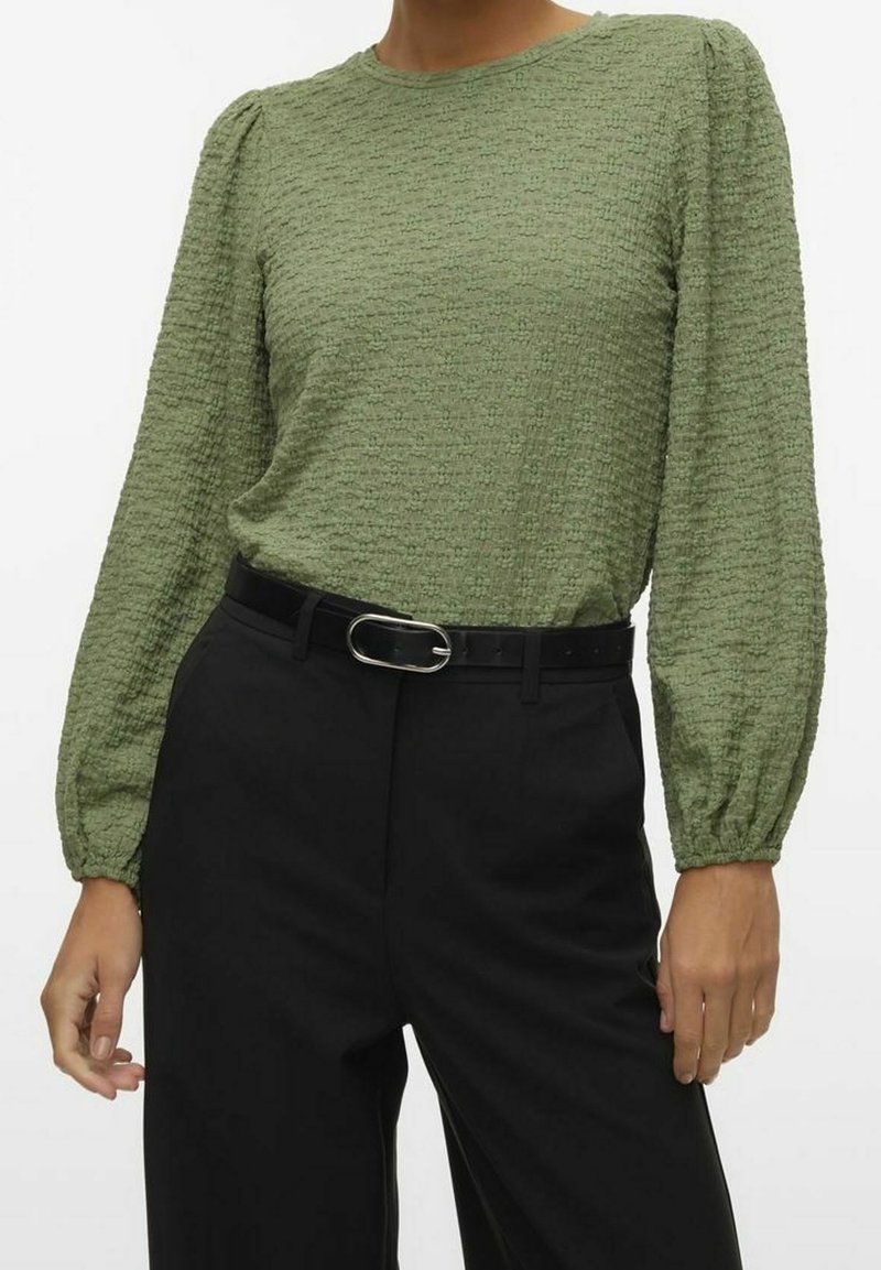 Femme portant un chemisier texturé vert olive à manches longues, rentré dans un pantalon noir taille haute avec une ceinture noire avec une boucle argentée.
