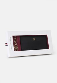 Svart läderplånbok med gold detaljer, med en logotyp och en nyckelring. Presenterad i en vit låda med röd inredning.