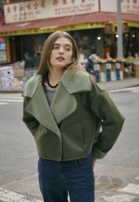 Olivewoy zieleń, krótka kurtka z pluszowym, teksturowanym kołnierzem, z podwójnym zapięciem i kieszeniami z przodu, w połączeniu z niebieskimi dżinsami.