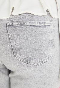 Pantalones de mezclilla gris con un bolsillo trasero con contorno cosido y una textura ligeramente desgastada. El dobladillo superior está parcialmente cubierto por una prenda.
