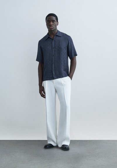 Άνδρας που στέκεται μπροστά σε ανοιχτόχρωμο τοίχο, φοράει μπλε ναυτικό κοντομάνικο πουκάμισο με κουμπιά, λευκό παντελόνι φαρδύ και μαύρα παπούτσια, με τα χέρια στις τσέπες.