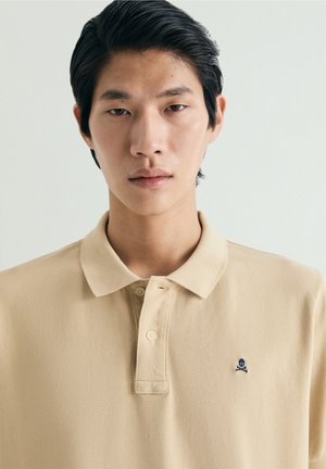 Ung mand med glat sort hår iført en beige polo-shirt med et lille mørkt logo af et kranium med krydsede knogler på brystet.