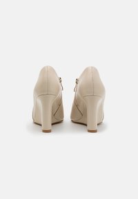 Beżowe szpilki z okrągłymi obcasami i bocznymi zamkami, widok z tyłu na jednolitym białym tle.
