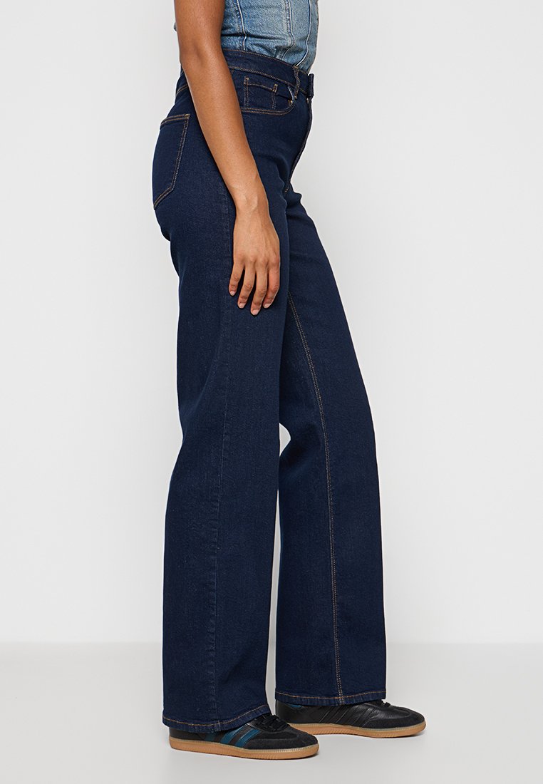 Dunkelblaue weite Jeans aus Denim, ausgestattet mit Seitentaschen, kontrastierenden Nähten und einem hoch sitzenden Design. Getragen mit schwarzen Schuhen.