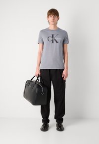 Gray short-sleeve t-shirt with "Calvin Klein Jeans" logo, black cargo pants, black leather duffel bag, and black shoes. Simple, clean design.