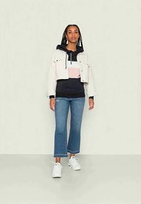 Jaqueta de ganga branca cortada sobre um hoodie preto com riscas rosa e creme, combinada com calças de flares azuis claras e ténis brancos.