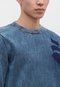 Jeune homme portant un pull tricoté bleu avec un col rond côtelé et un motif bleu foncé discret sur la poitrine.