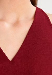 Zbliżenie szyi i klatki piersiowej osoby noszącej teksturowany, głęboko czerwony top na ramiączkach z dekoltem w kształcie litery V.